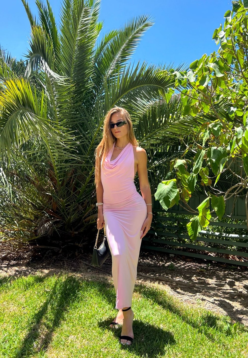 Femme portant une robe sans manches rose pâle, des talons noirs et des lunettes de soleil, debout sur l'herbe avec des palmiers et un feuillage vert en arrière-plan.