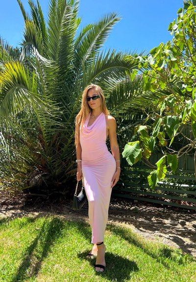 Mujer con vestido sin mangas rosa pálido, tacones negros y gafas de sol, de pie sobre el césped con palmeras y follaje verde en el fondo.