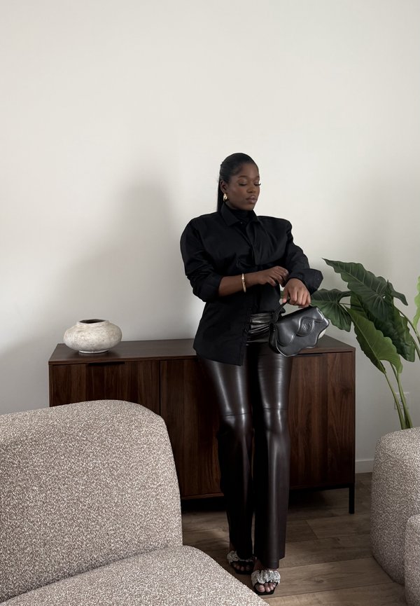 Femme en blouse noire et pantalon en cuir ajustant sa manche, tenant un sac à main noir, debout près d'une armoire en bois avec une plante et un vase à proximité.