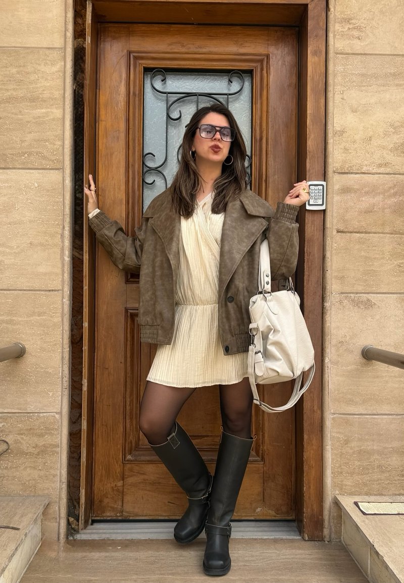 Ženska v preveliki rjavi jakni, kremni obleki, črnih škornjih in sončnih očalih stoji pred lesenimi vrati in drži belo ročno torbico.