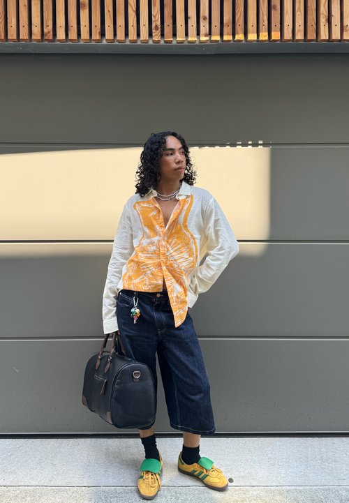 Avskuren vit t-shirt med grön kant, blå denim culotter och gula sneakers; bärande en keps och en mönstrad scarf; träd i bakgrunden.