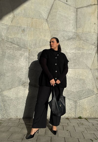 Chaleco negro de punto con mangas cortas y detalles de botones, combinado con pantalones negros y zapatos de tacón con punta. Un bolso negro completa el atuendo.