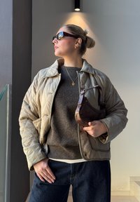 Casaco acolchoado beige, suéter cinza escuro, calças de ganga escuras, bolsa marrom na mão, óculos de sol tartaruga, acessórios em joias douradas.