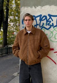 Hnědá semišová bomber bunda se zipem, elastickými manžetami a kapsami. Nošená přes bílou košili, v kombinaci s tmavými džínami. Městské pozadí.