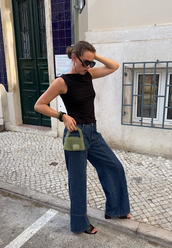 Femme avec un haut noir sans manches et un jean large posant dans une rue pavée, tenant un petit sac à main vert et portant des lunettes de soleil.