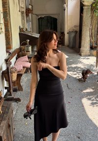 Schwarzes, tailliertes Kleid mit ausgestellt gesäumtem Saum. Glattes Material, Schultern freilassendes Design. Die Szene zeigt einen rustikalen Steinweg und Holzmöbel.