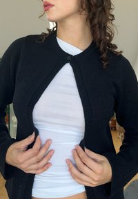 Μαύρο κασκόλ με στρογγυλή λαιμόκοψη, κλείσιμο με μονό κουμπί, ριγέ περιθώρια και μακριά μανίκια, φορεμένο πάνω από ένα λευκό μπλουζάκι.