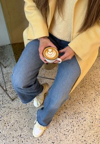 Baltas keraminis puodelis su latte menu, šviesiai ruda putų sūkuriais ant lygaus paviršiaus. Kasdienis šviesiai geltonas paltas, kreminis megztinis ir mėlyni džinsai.