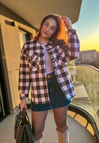 Černobílá kostkovaná košile, džínová sukně, hnědý pásek a béžové kozačky po kolena. Drží černou kabelku. Teplé světlo západu slunce.