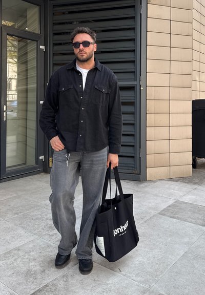 Hombre con camisa negra y gafas de sol está de pie afuera sosteniendo una bolsa tote negra con el texto "bonheur" cerca de la entrada del edificio.