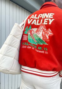 Casaco bomber de feltro vermelho com cintura de malha riscada branca, apresentando gráficos bordados de montanha verde e esquiador, texto "ALPINE VALLEY". Bolsa branca acolchoada.