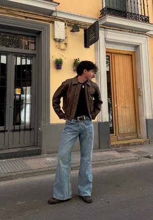 Hombre con chaqueta de cuero marrón y vaqueros anchos azules está de pie con las manos en las caderas en la calle junto a un edificio amarillo con puerta de madera y reja de metal.