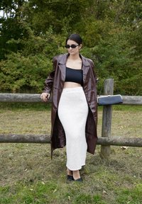 Brązowy błyszczący długi płaszcz, czarny top cropped, biały teksturowany maxi spódnica i czarne baleriny. Trzymając małą czarną torebkę przy drewnianym płocie.