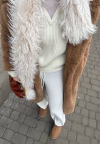 Άτομο που φοράει καφέ παλτό από Shearling με λευκό αφράτο γιακά, κρεμ πλεκτό πουλόβερ, λευκό παντελόνι, καφέ μπότες χωρίς κορδόνια και κρατάει καφέ δερμάτινη τσάντα.
