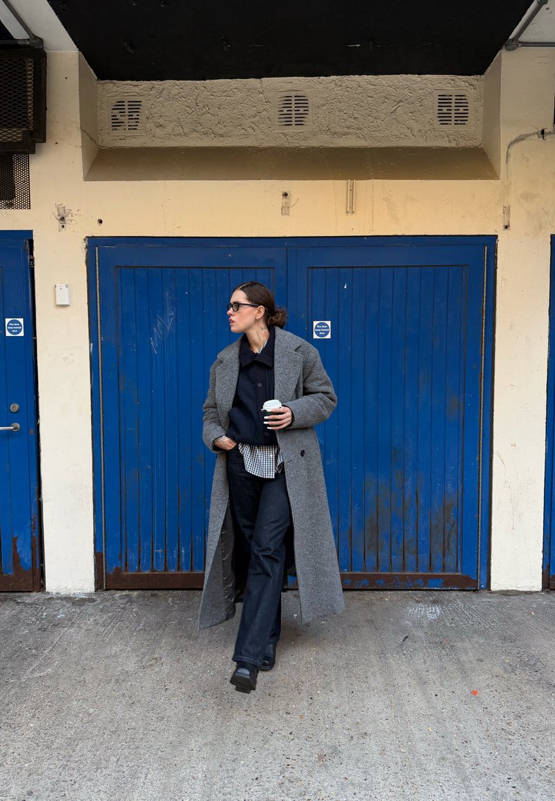 Femme en long manteau gris, pantalon noir et lunettes de soleil marchant devant des portes de garage bleues, tenant une tasse de café dans sa main droite.