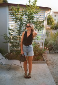 Black sleeveless button-up top, blue frayed denim shorts, black sandals with gold accents, holding a small black woven bag; outdoor setting.