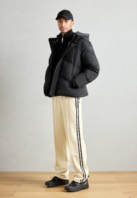 Sort polstret jakke med hætte, lynlåslukning og logo på brystet. Cremefarvede bukser med sorte sidestreger, kombineret med sorte sneakers.