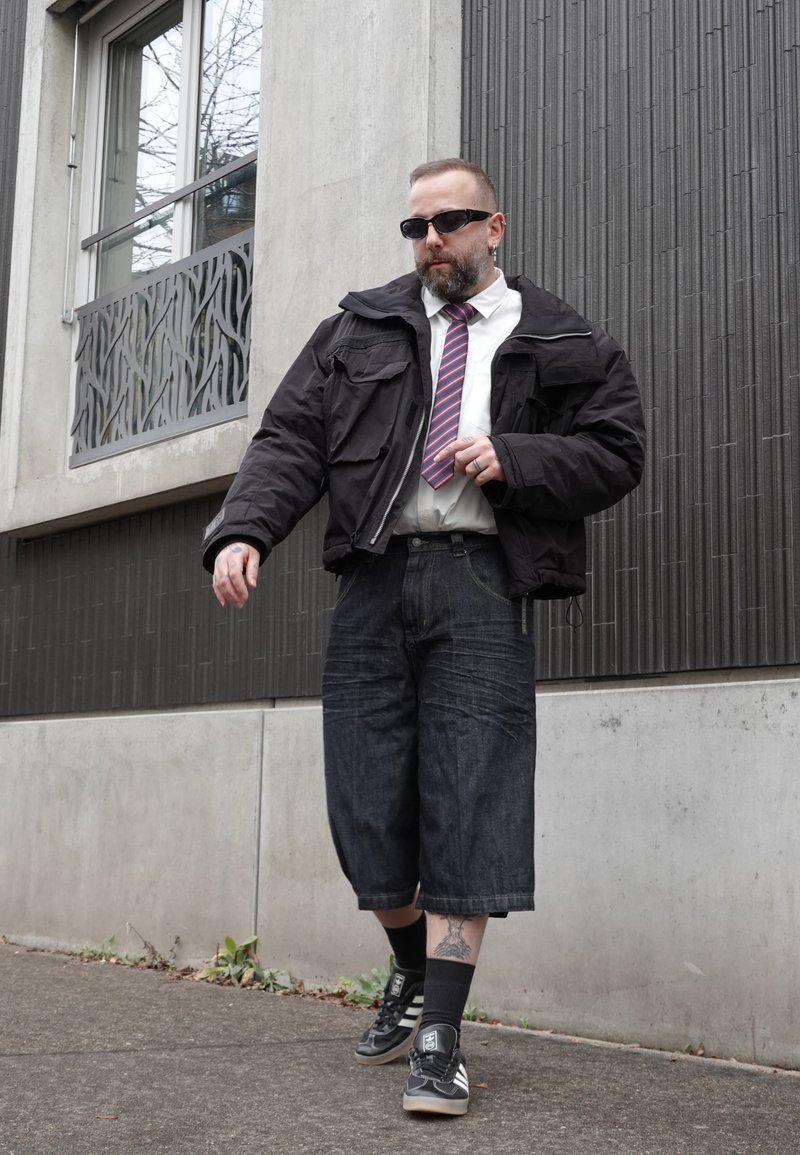 Hombre con gafas de sol, chaqueta negra, corbata a rayas, pantalones cortos de mezclilla largos y zapatillas caminando por la acera de una ciudad junto a un edificio de concreto.