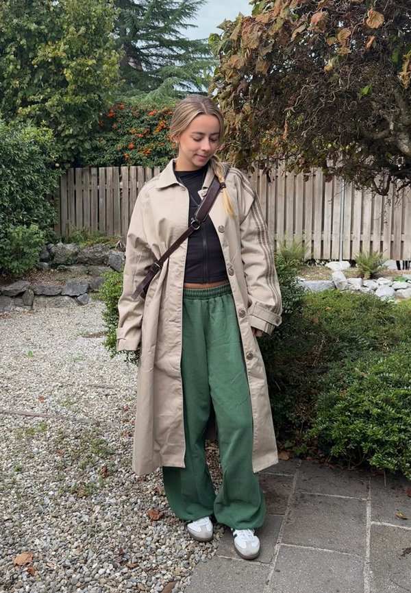 Joven mujer está al aire libre vistiendo un abrigo largo beige, pantalones verdes, zapatillas blancas y un bolso cruzado, con árboles y una valla de madera detrás de ella.