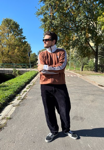 Chaqueta de chándal marrón con cremallera y cuello blanco, combinada con pantalones de pana negros y zapatillas en blanco y negro en un sendero.