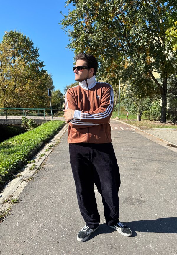 Giacca da track nera con zip, colletto bianco e tre strisce bianche, abbinata a pantaloni in velluto a coste neri e sneaker bianco e nere su un sentiero.