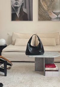 Sac à main en cuir noir sur une table basse en marbre devant un canapé beige avec des coussins, pile de livres sous la table, et deux portraits muraux au-dessus.