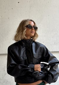 Person med blondt bølget hår, iført svarte solbriller og en svart skinnjakke, holder en svart teksturert håndveske foran en betongvegg.