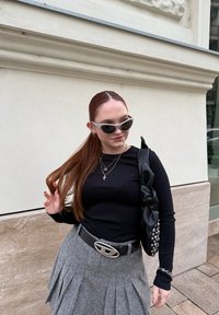 Svart långärmad topp, grå plisserad kjol, silvrigt bältesspänne, texturerad svart handväska med rosett och nitar, klara solglasögon, rakt brunt hår.