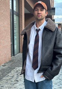 Jaqueta de piloto em pele castanha com gola de pelagem, camisa branca e gravata de riscas escuras. Jeans azuis casuais e boné bege. Ambiente urbano.