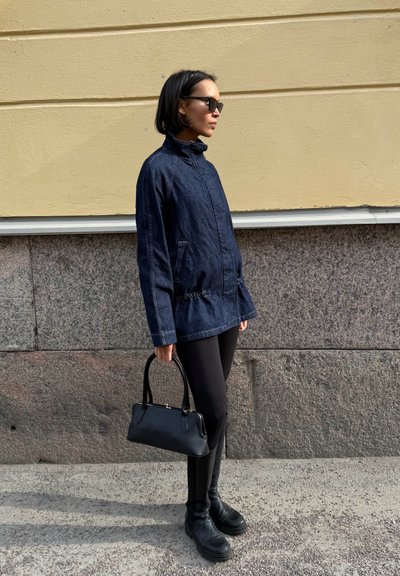 Mujer con gafas de sol negras, chaqueta de mezclilla oscura, leggings negros y botas, sosteniendo un bolso negro, de pie frente a una pared de piedra y beige.