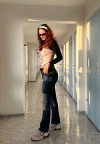 Lilla crop top med lace-detaljer, sort langærmet skjorte, blå jeans med slidte pletter, hvide sneakers og lilla solbriller i en korridor.