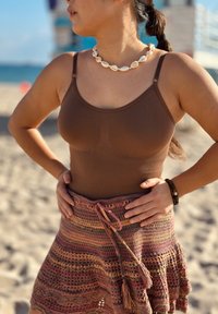 Femme debout sur une plage de sable, portant un débardeur marron, une jupe en crochet multicolore, un collier de coquillages et un bracelet, les mains sur les hanches.