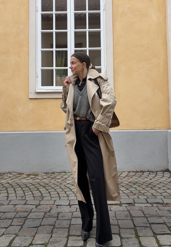 Una mujer con una gabardina beige y pantalones negros está de pie en una calle empedrada junto a un edificio amarillo y mira hacia un lado.