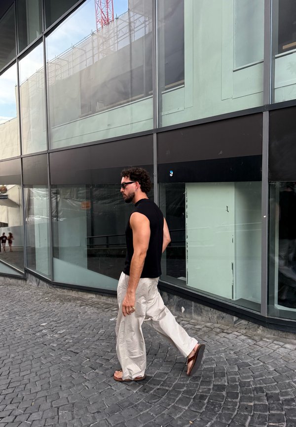 Uomo con maglietta nera senza maniche, pantaloni beige, sandali e occhiali da sole che cammina su una strada acciottolata accanto a un edificio di vetro.