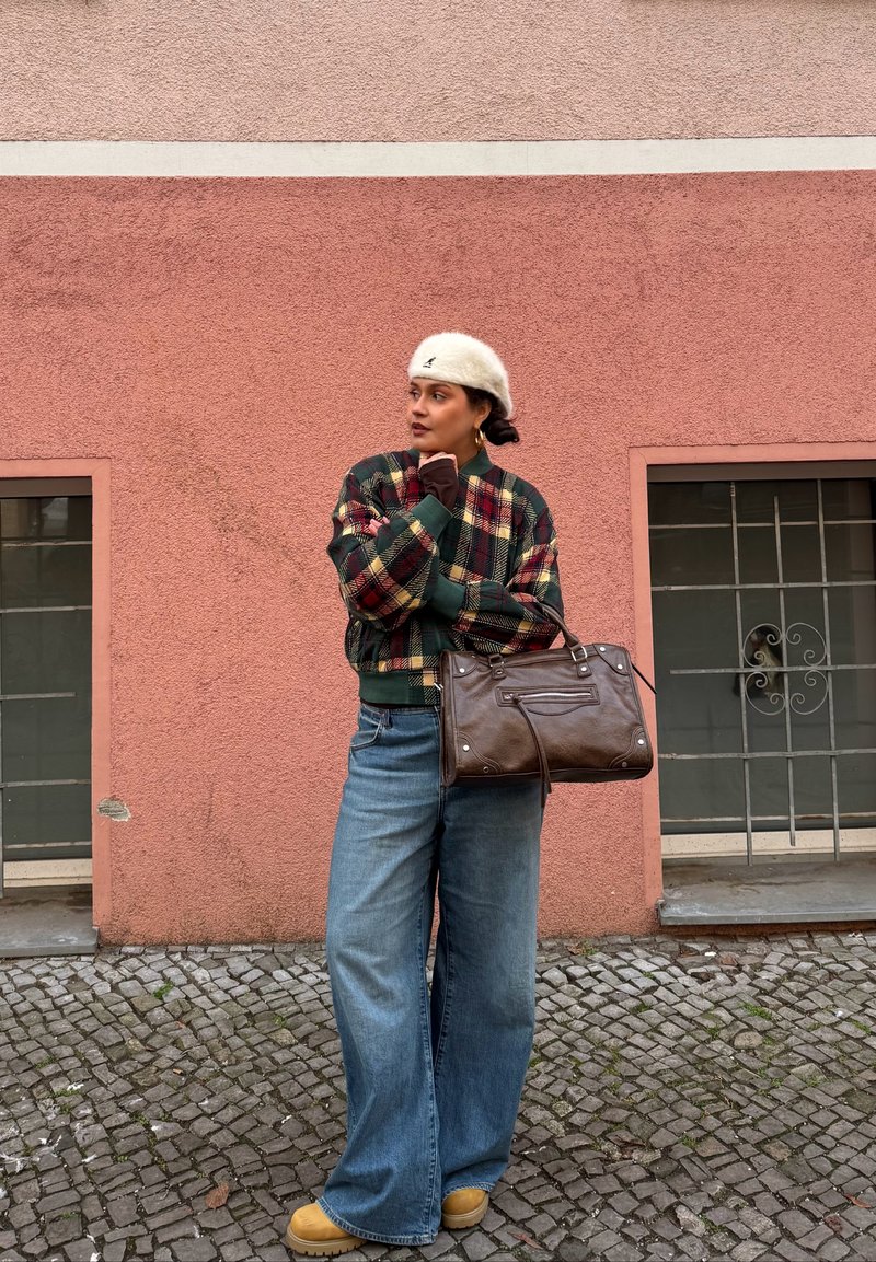 Frau mit weißem Barett, kariertem Jackett, weitem Jeans und gelben Stiefeln, die eine braune Handtasche hält und auf einem Kopfsteinpflaster vor einer rosa Wand steht.
