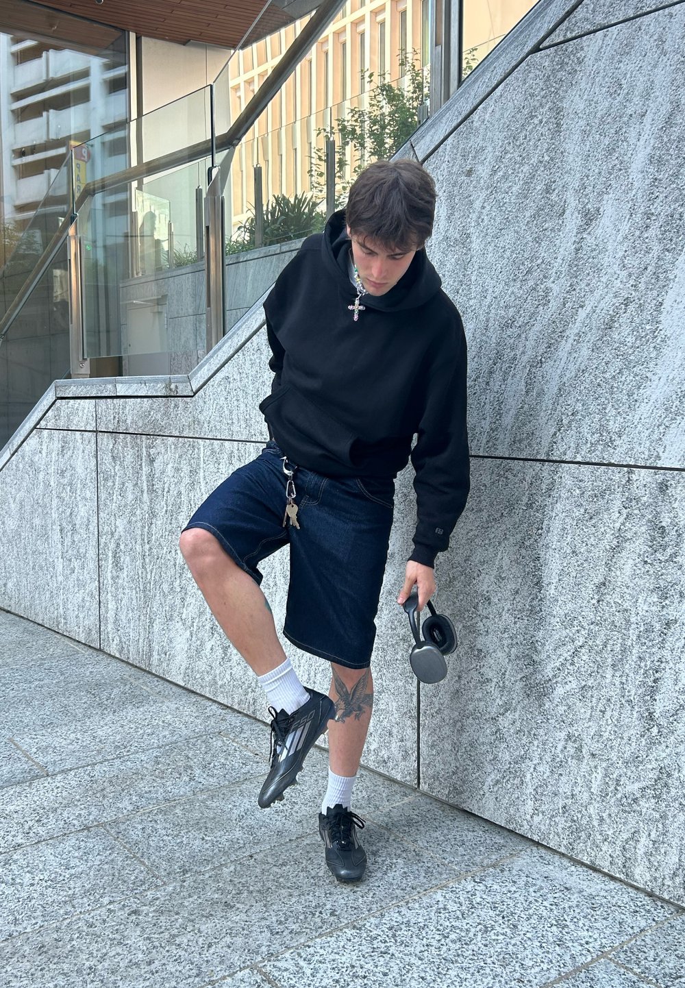 Schwarzer Kapuzenpullover, Denim-Shorts, schwarze Sneaker; Kopfhörer haltend, auf Steinstufen stehend mit einer Betonmauer im Hintergrund.