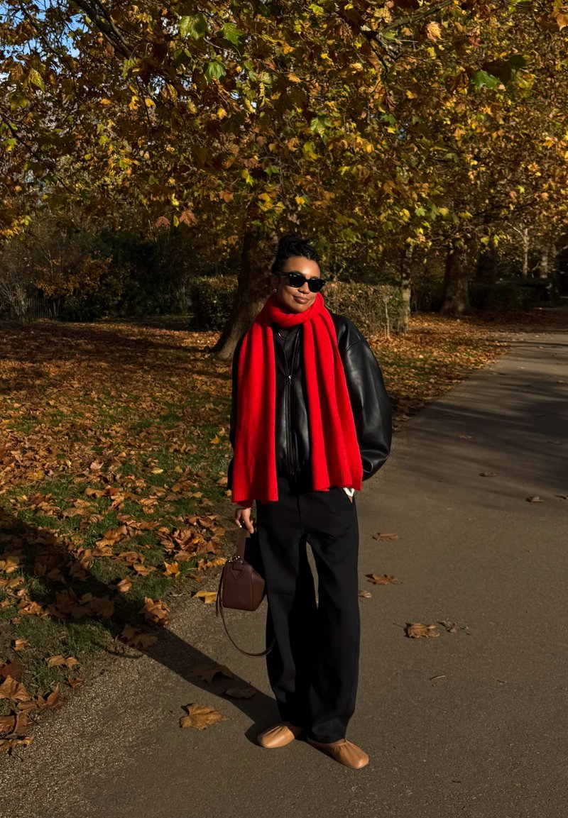 Femme portant une veste en cuir noire et une écharpe rouge vif, debout sur un chemin couvert de feuilles dans un parc d'automne, tenant un sac à main marron.