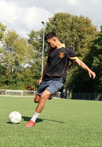 Mladý muž v černém a oranžovém sportovním oblečení, který kopne bílý fotbalový míč na zeleném venkovním hřišti s stromy v pozadí.