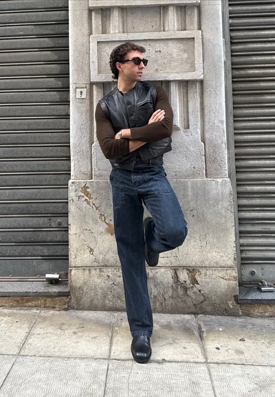 Hombre con gafas de sol, chaleco de cuero y jeans se apoya contra una pared de piedra con los brazos cruzados y un pie levantado sobre la acera.