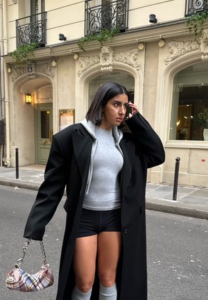 Joven mujer con abrigo largo negro, suéter gris con capucha, pantalones cortos negros y calentadores de piernas de punto gris, sosteniendo un bolso de cuadros en una calle de la ciudad.