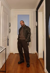 Ciemnobrązowa skórzana kurtka na brązowym swetrze i dopasowanych spodniach; połyskujące czarne buty. Jasna drewniana podłoga i białe drzwi w tle.