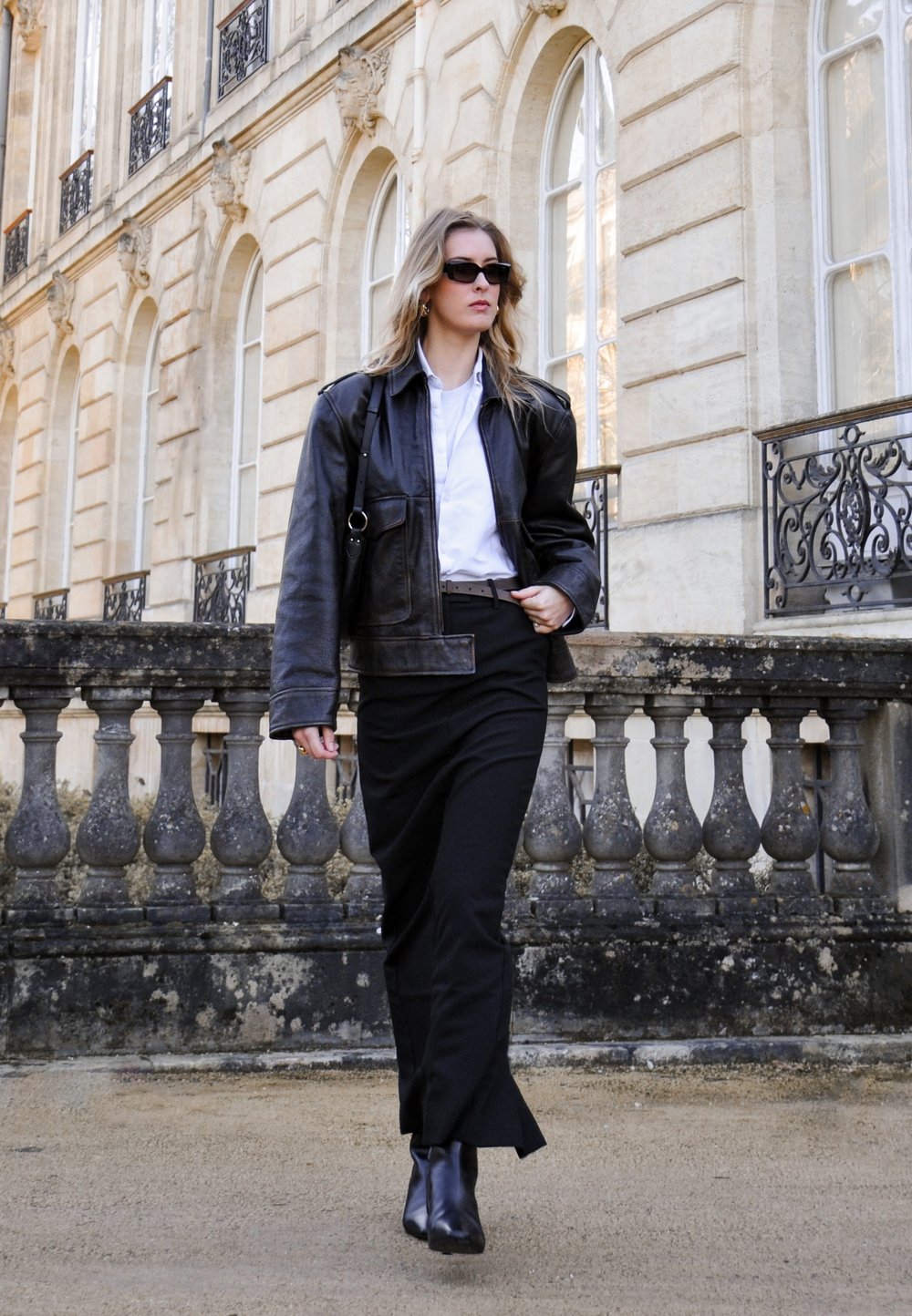 Black leather jacket over a white shirt, paired with a black skirt and ankle boots. Setting features classic architecture and stone balustrade.