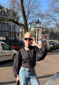 Črni skrajšan top s puff rokavi, v kombinaciji s svetlo modrimi jeans hlačami. Dodatki vključujejo črni ovratnik in sončna očala. Urbano ozadje.