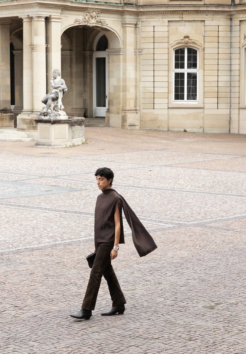 Person in a dark brown outfit with a flowing scarf walks on a cobblestone courtyard near a stone sculpture and a historic building with arches and windows.