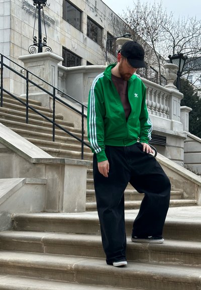 Chaqueta verde con cremallera y rayas blancas, pantalones negros holgados y zapatillas negras, de pie en unos escalones de piedra fuera de un edificio.