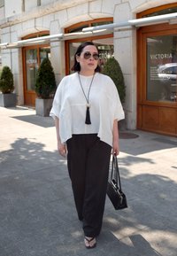 Top blanco de gran tamaño combinado con pantalones negros de pierna ancha. Los accesorios incluyen un collar largo con borlas y un bolso negro. Ambiente soleado al aire libre.