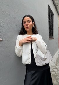 Valge, pehme kardigan nööpidega, kantud musta topi ja seelikuga. Hoides hõbedast võrgust kotti dekoratiivse rihmaga. Lokkis juuksed.