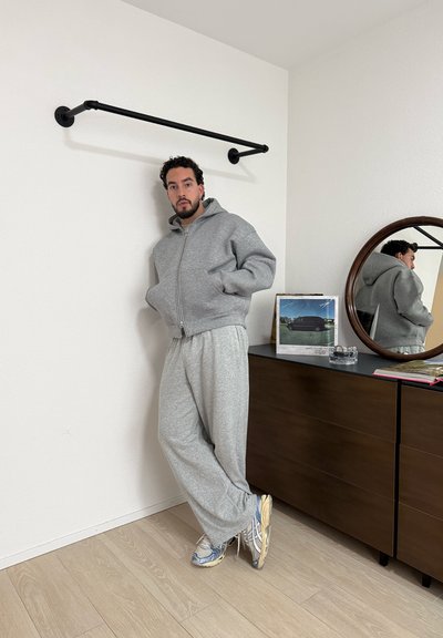 Hombre con sudadera gris con capucha y pantalones deportivos apoyado contra una pared blanca cerca de una cómoda oscura con espejo redondo que refleja su perfil.