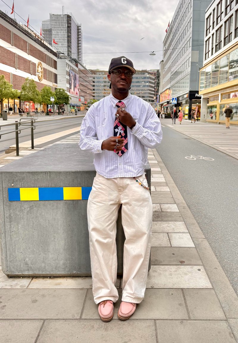 Homme portant une chemise blanche, une cravate à motifs, un pantalon blanc, des chaussures roses et une casquette noire, se tient sur le trottoir de la ville à côté d'un bloc de béton avec des rayures bleues et jaunes.