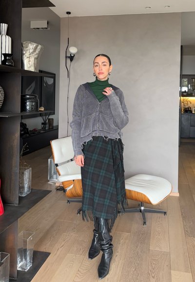 Mujer con cabello liso que lleva un suéter verde de cuello alto, un cárdigan gris con cremallera, una falda de cuadros con flecos y botas negras, de pie en una habitación moderna con una silla.